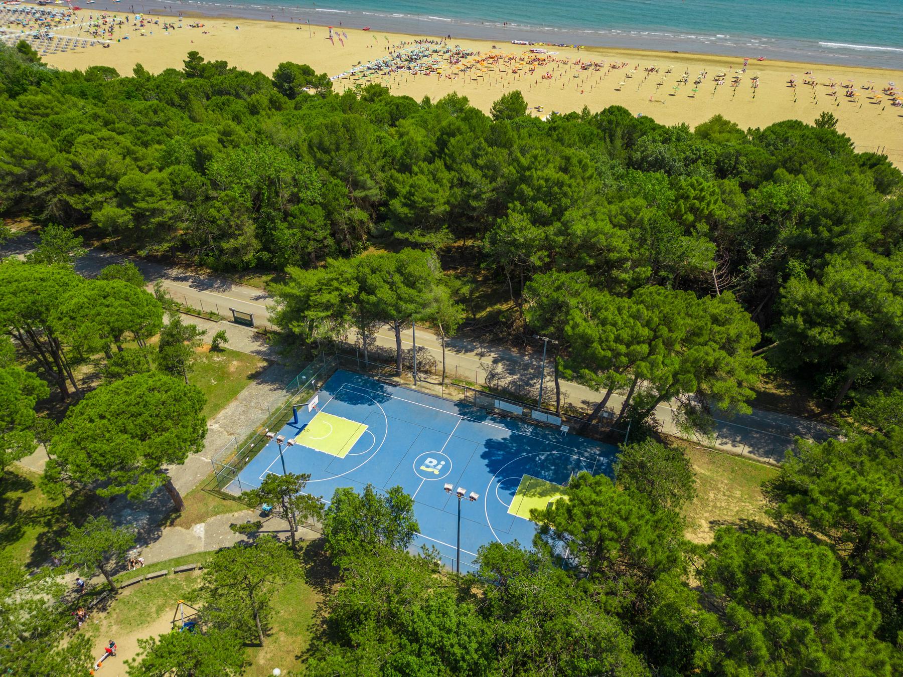 Luftbild: Basketball- und Volleyballfeld im Pinienwald, dahinter der Strand und das Meer