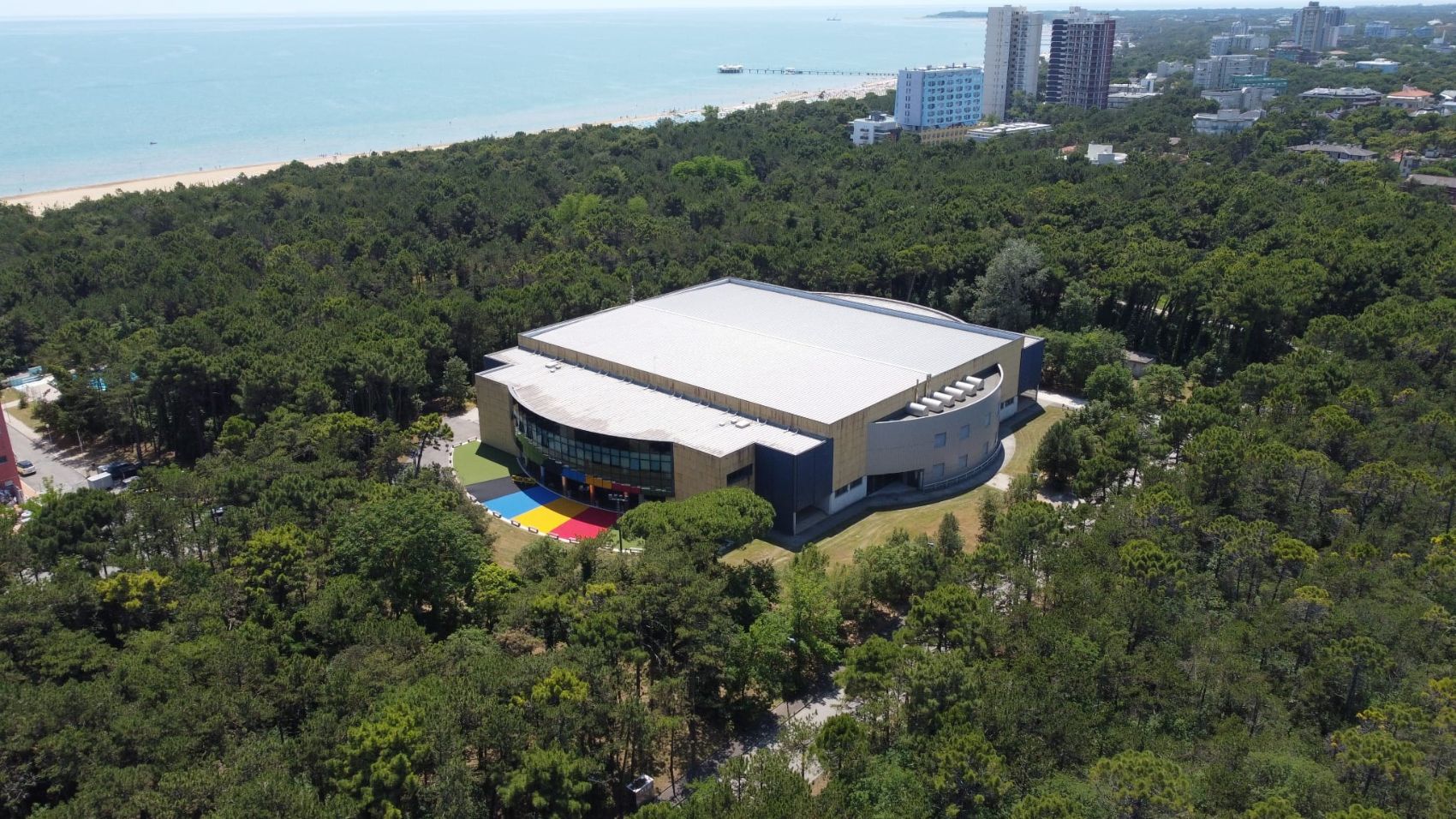 Luftbild der Sporthalle im Pinienwald von Lignano, dahinter das Meer