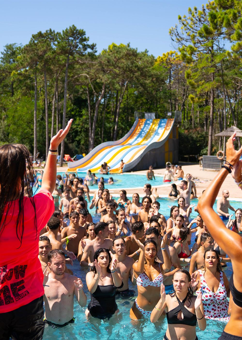 Poolparty im Wasserpark der Anlage: Jugendliche tanzen im Becken, dahinter die bunte Breitrutsche im Pinienwald