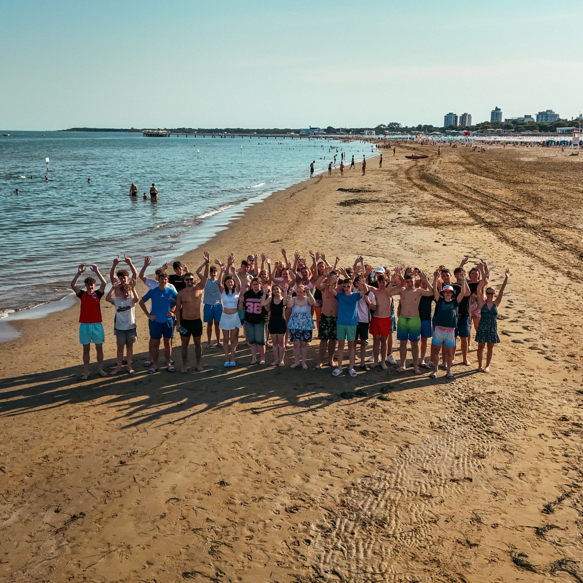 Schulklasse am Strand von Lignano, die Jugendlichen winken mit erhobenen Armen in die Kamera